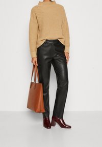 Suéter beige de punto, pantalones de cuero negro, botas altas burdeos y un bolso tote marrón. Tela texturizada con un ajuste relajado y cuello alto.