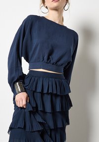 Marineblaue, kurz geschnittene Bluse mit weiten Ärmeln und gerafftem Saum, kombiniert mit einem marineblauen Faltenrock mit horizontalen Rüschen und strukturierter Stoffqualität.