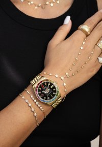 Goldene Uhr mit schwarzem Zifferblatt und mehrfarbigen Stundenmarkierungen, begleitet von goldenen Armbändern und Ringen mit kleinen dekorativen Steinen.