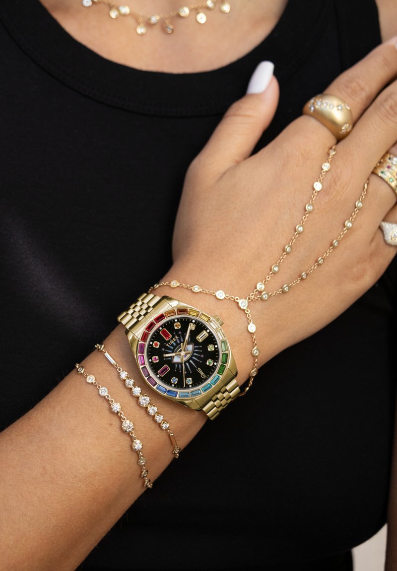 Goldene Uhr mit schwarzem Zifferblatt und mehrfarbigen Stundenmarkierungen, begleitet von goldenen Armbändern und Ringen mit kleinen dekorativen Steinen.