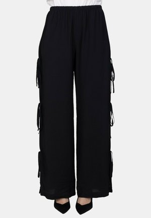 Pantaloni neri a gamba larga con vita elastica e lacci decorativi sui lati, indossati con scarpe nere con punta a punta.