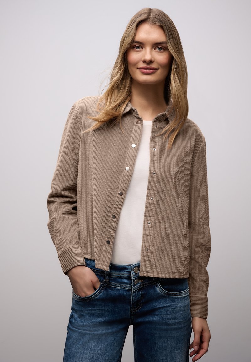 Jeune femme aux longs cheveux blonds portant une veste en velours côtelé beige sur une chemise blanche et un jean bleu, debout avec une main dans la poche.