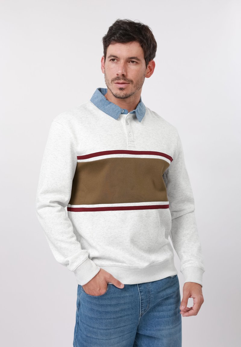 Homme portant un pull blanc avec des rayures marron et bordeaux et un col bleu, debout avec une main dans la poche sur un fond uni.