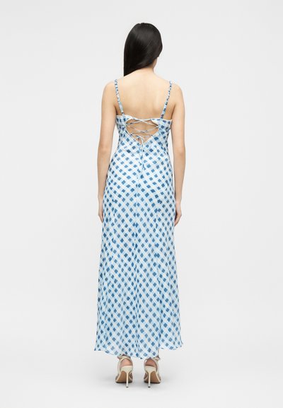 Γυναίκα με μακριά σκούρα μαλλιά, που φοράει μακρύ φορεματάκι χωρίς μανίκια, μπλε και λευκό καρό, με δέσιμο στο πίσω μέρος και μπεζ παπούτσια με ψηλό τακούνι.