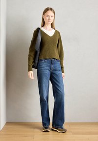 Olivengrønn ribbestrikket genser med V-hals, kombinert med blå vide jeans og grå sneakers, holder en svart veske.