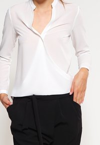 Bílá, lehká blůza s výstřihem do V a dlouhými rukávy; má zavinovací design a jemný lesk; kombinována s černými kalhotami na šňůrku.