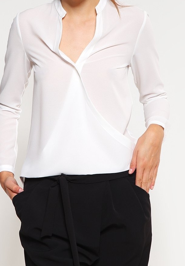 Bílá, lehká blůza s výstřihem do V a dlouhými rukávy; má zavinovací design a jemný lesk; kombinována s černými kalhotami na šňůrku.