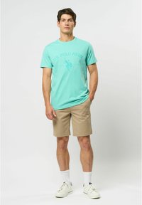 Tricou teal deschis cu logo-ul U.S. Polo Assn., asociat cu pantaloni șort bej. Modelul poartă șosete albe și teneși. Design simplu și casual.