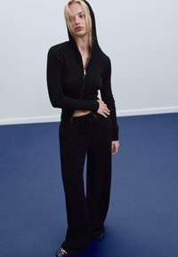 Schwarze, gerippte Zip-Up-Hoodie mit Kapuze, kombiniert mit lässigen, weiten schwarzen Jogginghosen. Das Outfit zeichnet sich durch ein minimalistisches Design und eine lockere Passform aus.