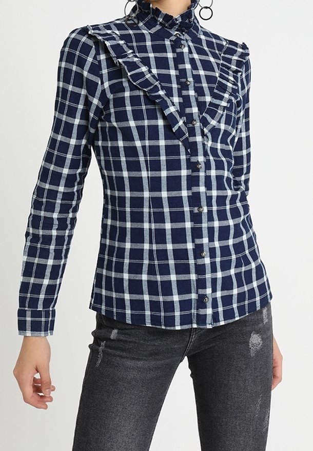 Chemise à carreaux bleu marine et blanche à boutons avec des détails à volants sur la poitrine, portée avec un jean noir déchiré.