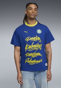 Blå bomulds t-shirt med fed gul skrift af "Palmeiras" og et rundt logo på venstre bryst, suppleret af en kontrasterende gul krave.