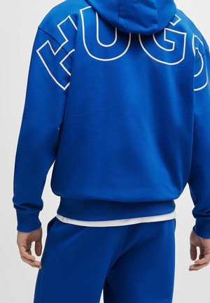 Man draagt een blauw hoodie en bijpassende blauwe broek, met witte omlijnde letters op de achterkant van het hoodie, staand tegen een effen achtergrond.