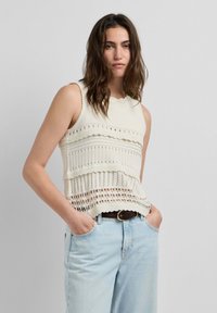 Frau mit welligem braunem Haar, die ein ärmelloses weißes Strickoberteil mit Mustern und hellblaue Jeans trägt, mit den Händen in den Taschen stehend.