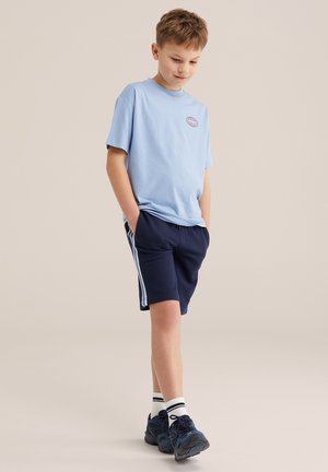 Băiat tânăr cu tricou bleu deschis și pantaloni scurți bleumarin cu dungi laterale, cu mâinile în buzunare, privind în jos, stând pe un fundal bej.