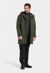 Olijfgroene waterdichte jas met capuchon, knoopsluiting aan de voorkant en twee zijzakken, gedragen over een zwart gevoerd jack en donkere broek.