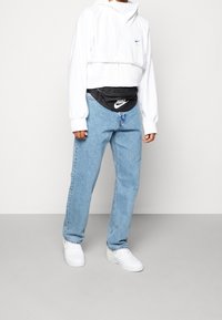 Valkoinen lyhyt huppari, jossa on narut, vaaleansiniset farkut, musta Nike-vyölaukku ja valkoiset lenkkarit. Siisti, rento katutyylinen asu.