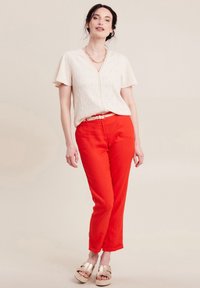 Weiße Bluse aus Lochstickerei mit kurzen Ärmeln, kombiniert mit knallroten, geraden Hosen und goldenen Sandalen. Besitzt einen Gürtel mit einer Schnallenakzentuierung.