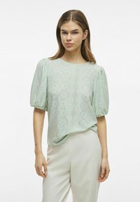 Blusa verde chiaro con maniche a palloncino, caratterizzata da intricate lavorazioni a occhio di pernice. Texture del tessuto morbida, scollatura rotonda e vestibilità comoda.