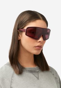 Prevelika bordo sončna očala z geometrijskim dizajnom, temno obarvanimi lečami in tankimi kraki, ki dopolnjujejo priložnostno sivo jopa.