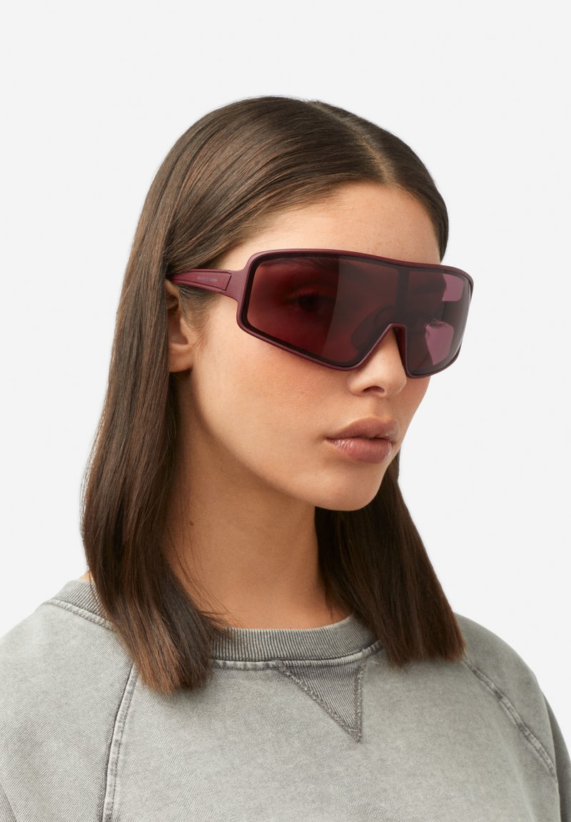 Prevelika bordo sončna očala z geometrijskim dizajnom, temno obarvanimi lečami in tankimi kraki, ki dopolnjujejo priložnostno sivo jopa.