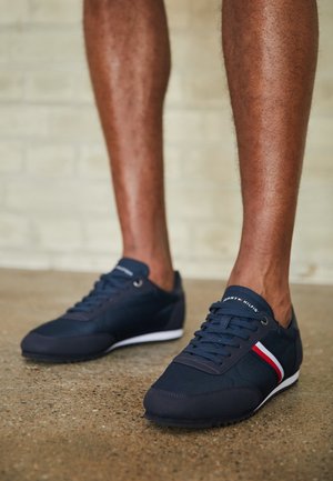 Nogi w granatowych tenisówkach Tommy Hilfiger z białymi i czerwonymi bocznymi paskami, stojące na teksturowanej betonowej podłodze.