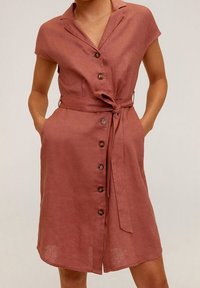 Abito di lino color rosa con maniche corte, chiusura a bottoni e cintura annodata in vita. Presenta due tasche frontali e una lunghezza fino al ginocchio.