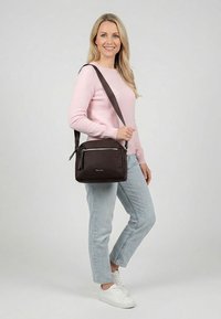 Smaidoša sieviete rozā džemperī, gaišās džinsās un baltos sporta apavos nēsā melnu plecu somu ar rāvējslēdzēja kabatām.