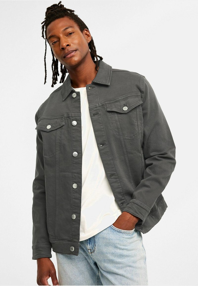 Grå denimjacka med krage, frontknäppning, två bröstfickor och långa ärmar. Burken över en vit T-shirt och ljusblå jeans.