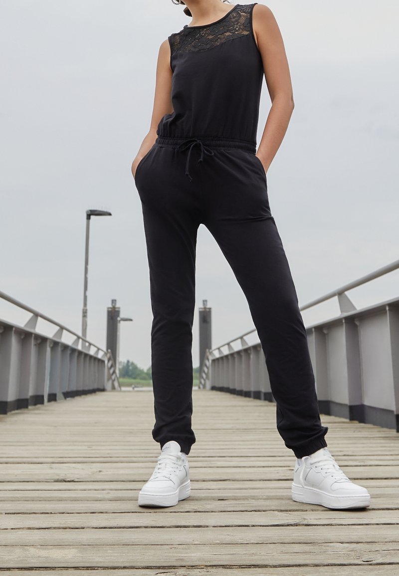 Femme debout sur une jetée en bois portant une combinaison noire sans manches avec un détail en dentelle et des baskets blanches, les mains dans les poches.