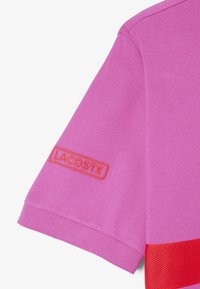 Rosa polo-skjorta med texturerad yta, med en orange broderad "LACOSTE"-logga på ärmen och en röd detalj vid fållen.