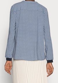 Persona che indossa una blusa blu a maniche lunghe con motivo e polsini neri, insieme a una gonna plissettata bianca, vista da dietro.