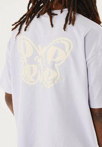 Camiseta de color púrpura claro con un gran gráfico estilizado de una mariposa blanca en la parte posterior y texto en cursiva, con mangas cortas y un ajuste relajado.