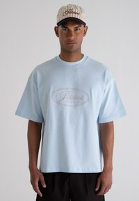Light blue oversized t-shirt made of cotton, featuring a central embroidered logo in white and pink in an oval design. Worn with a cap.