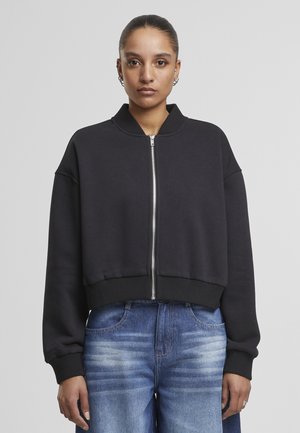 Čierna skrátená bomber bunda vyrobená z mäkkej tkaniny, s uzatváraním na zips, rebrovaným lemom a širokými rukávmi. Nosená cez modré roztrhnuté džínsy.