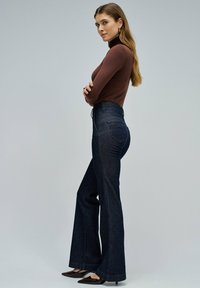 Jeans de mezclilla de tiro alto y acampanados en azul oscuro, combinados con un jersey ajustado de cuello alto marrón. Textura suave y detalles de costura sutiles.