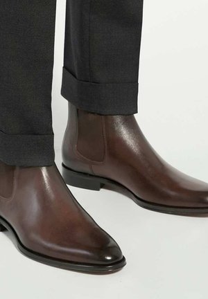 Brown leather chelsea boots with a glossy finish, pointed toes, and elastic side panels, paired with dark tailored pants.