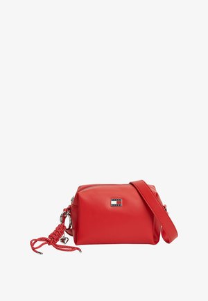 Bolso bandolera rojo de forma rectangular con cierre de cremallera, logo de Tommy Jeans en la parte frontal y cuerda trenzada roja con charm de corazón plateado adjunto.