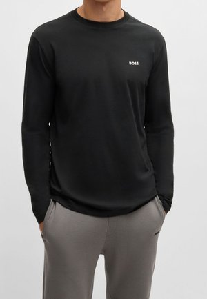 Homme portant un t-shirt noir à manches longues avec le logo "BOSS" sur la poitrine et un pantalon gris, les mains dans les poches, sur un fond uni.