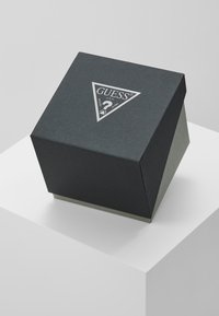 Svart texturerad presentask med ett triangulärt lock, med silverfärgad GUESS-logotyp på toppen, vilande på en vit pelare.