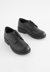 Chaussures en cuir noir avec un bout arrondi, un talon bas et un design à lacets. Semelle texturée avec un motif de crampon visible pour une meilleure adhérence.