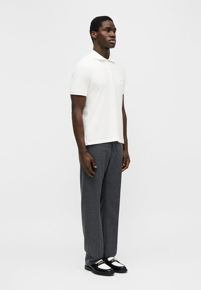 Homme debout portant un polo blanc à manches courtes, un pantalon gris et des mocassins noirs et blancs, devant un fond clair uni.