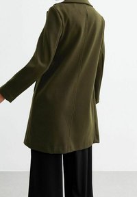 Olivgrön kappa med krage, mjuk textur, långa ärmar och figursydd passform, bärs över svarta byxor. Bakifrån syns enkla designlinjer.