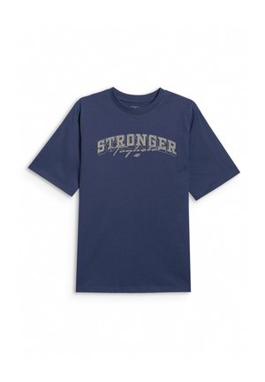Chłopiec stojący, ubrany w granatowy luźny T-shirt z napisem „STRONGER”, szare spodnie dresowe z nadrukiem po bokach oraz szare trampki z różowymi sznurowadłami.