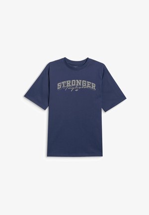 Chłopiec stojący, ubrany w granatowy luźny T-shirt z napisem „STRONGER”, szare spodnie dresowe z nadrukiem po bokach oraz szare trampki z różowymi sznurowadłami.