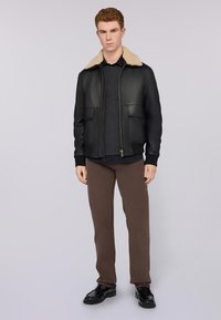 Giubbotto bomber in pelle nera con colletto in pelliccia color cioccolato, due tasche frontali, polsini a costine, indossato su un maglione scuro e pantaloni marroni.