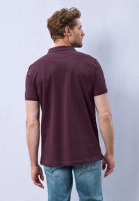 Polo à manches courtes en tissu bordeaux, avec un col classique, des ouvertures latérales, et une coupe décontractée, associé à un jean en denim bleu clair.