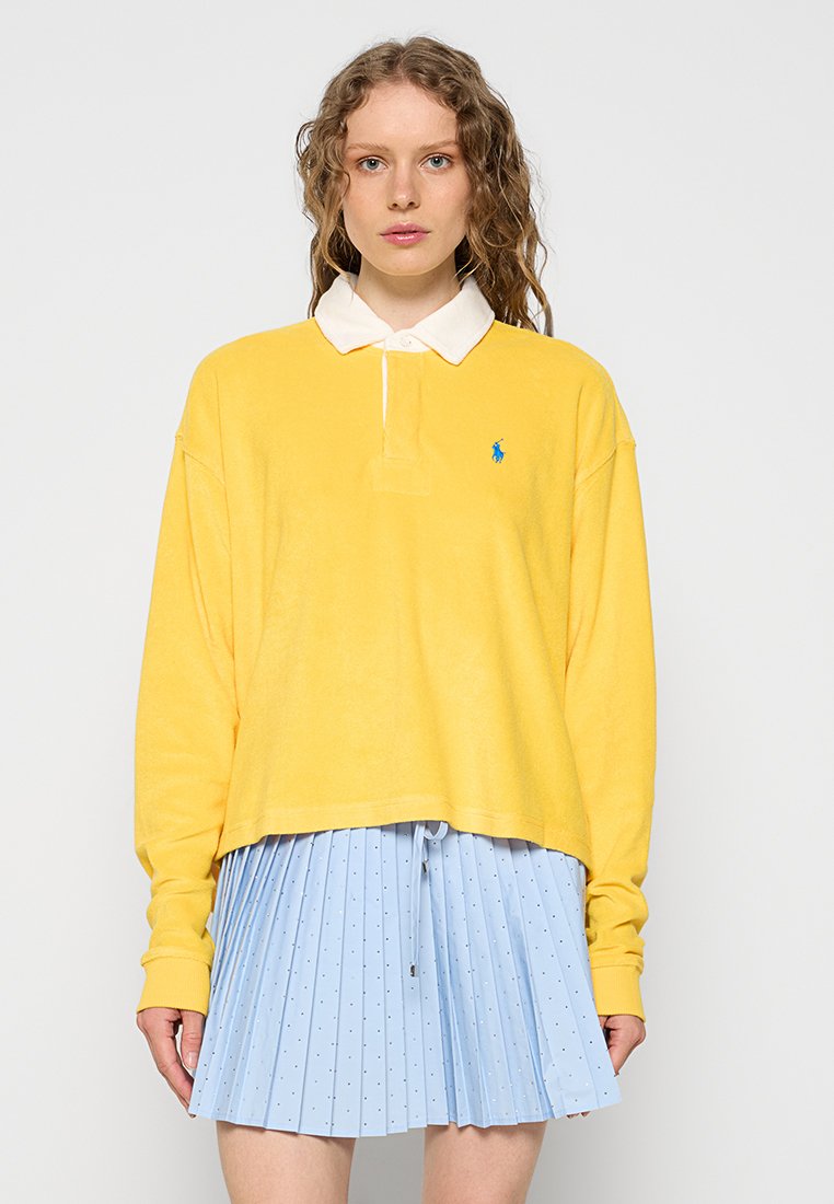 Polo giallo a maniche lunghe con colletto bianco e logo blu sul petto, abbinato a una gonna plissettata azzurro chiaro con piccoli pois.