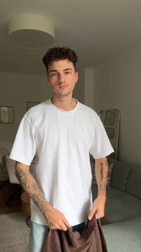 Camiseta blanca oversize de algodón, con cuello redondo, mangas cortas, con intrincados tatuajes en los brazos; sosteniendo una prenda de color marrón oscuro. Fondo neutro.