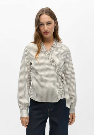 Femme aux longs cheveux bruns portant un chemisier blanc à rayures verticales marron avec un détail drapé à volants, associé à un jean bleu foncé.