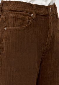Gros plan sur un pantalon en velours côtelé marron montrant la poche avant, le bouton et les passants de ceinture sur un fond de chemise blanche.